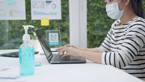 女性在使用笔记本电脑工作前应戴上口罩和用酒精凝胶消毒手冠状病毒危机后的新常态、社交距离措施和工作生活方式