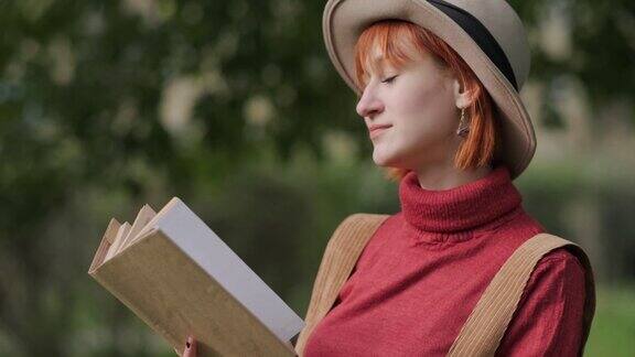 年轻迷人的红头发女人戴着帽子和毛衣在自然公园看书秋天凉爽的一天