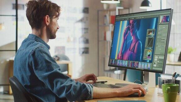穿着牛仔裤衬衫的男性数码编辑在他的大屏幕个人电脑上使用照片编辑软件他在很酷的办公室阁楼工作其他有创意的女性同事走在后面