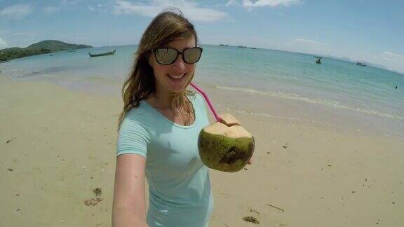 自拍:微笑的女游客漫步在沙滩上用椰子喝饮料