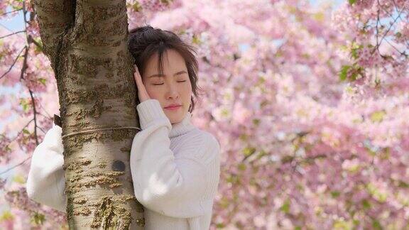 美丽的年轻女子抱着树干对着粉红色的樱花树享受风吹她的脸和头发闭上眼睛4k镜头慢镜头