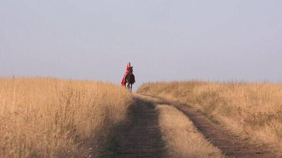 年轻的女孩穿着深红色的长裙骑着黑马穿过干燥的草原