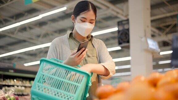 亚洲妇女戴着口罩拿着购物篮在超市COVID-19疫情期间女性在杂货店货架上购物和查看水果