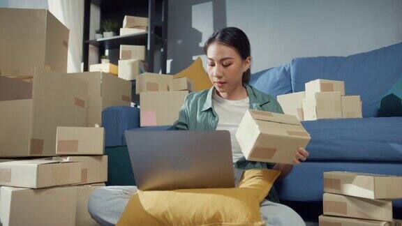 年轻的亚洲女售货员在网上商店使用笔记本电脑准备包裹发货给客户在家里