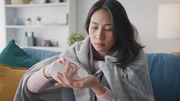生病的亚洲妇女喝水喝处方药在家里客厅的沙发上盖着毯子忍受着头痛或抑郁