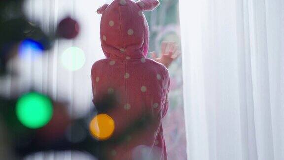 后视图快乐的孩子挥舞着看着窗外模糊的圣诞灯在室内前面中景轻松的白人女孩在家里享受新年