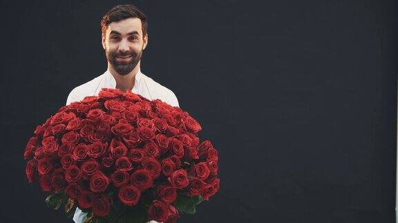 迷人的男子站在那里捧着一大束红玫瑰扬起眉毛面带微笑躲在花丛后面