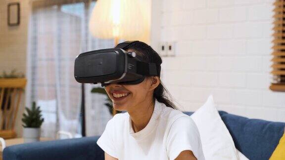 亚洲女性戴着vr头盔眼镜坐在家里的沙发上玩虚拟现实数字tehcnology生活方式..