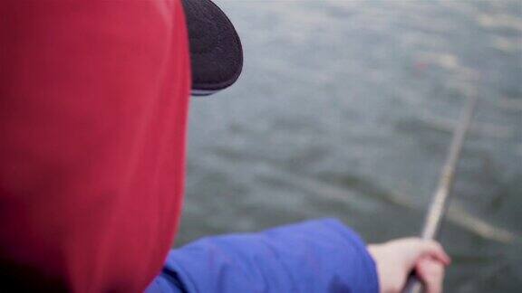 近距离的男孩钓鱼在河在帽子和兜帽和蓝色夹克