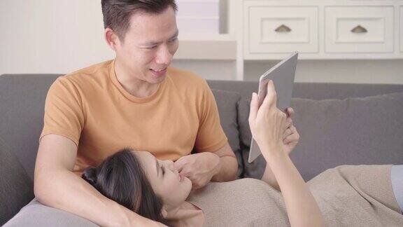 亚洲情侣在家中客厅用平板电脑上网购物甜蜜情侣在家中放松时躺在沙发上享受爱情时刻生活方式夫妇在家里放松