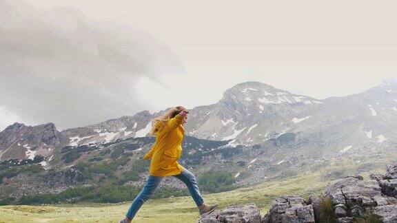 年轻快乐的女人穿着黄色的雨衣在美丽的山脉背景的岩石上奔跑慢镜头