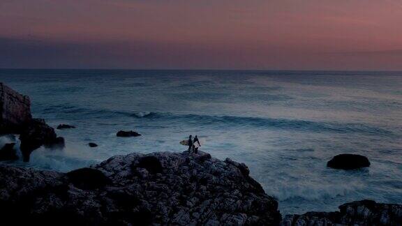 惊人的航拍照片两个女性冲浪者站在悬崖岩石的边缘观看日出时的海浪
