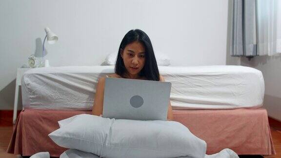 年轻的亚洲女人使用笔记本电脑查看社交媒体感到幸福的微笑而躺在床上早上在家里醒来后迷人的泰国女孩微笑放松在卧室在家里
