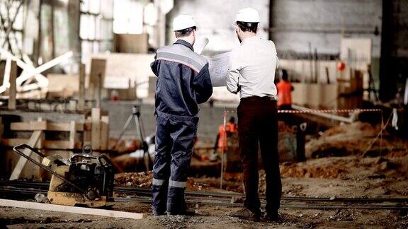 两个人站在一起的建筑区域和看未来的结构施工图的后视图穿衬衫和裤子的工头正在和工人讨论堆垛的细节