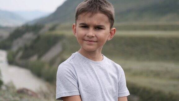 肖像穿着灰色t恤的小游客男孩看着相机背景是山脉和河流