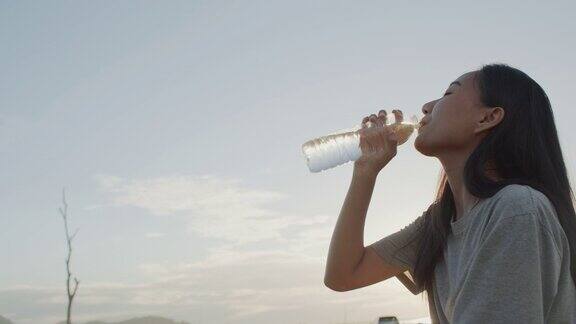 亚洲女孩坐在自然中喝瓶装水当阳光日落在周末时间
