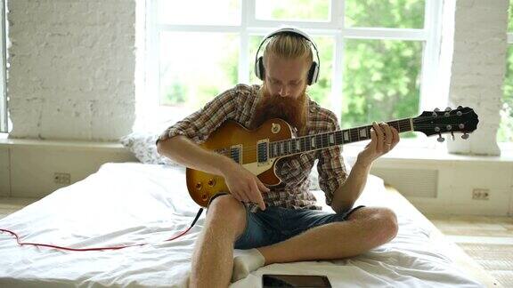 有魅力的大胡子男子坐在床上学习弹吉他在家里的现代卧室里用平板电脑