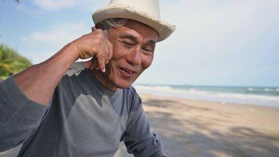 亚洲老人戴着帽子坐在沙滩上微笑着用手势打电话聊天