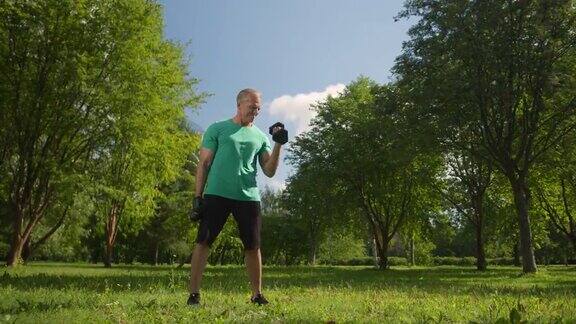 一个夏天的早晨一个身穿绿松石t恤的金发健美男子在公园拿着哑铃锻炼身体