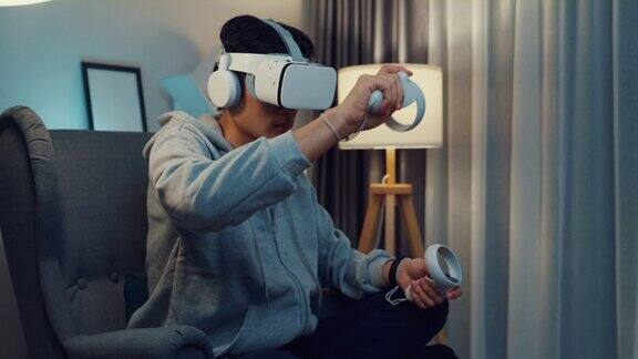 年轻的亚洲男子玩家穿着灰色卫衣与VR控制器头戴手柄操纵杆控制器创造3D艺术坐在沙发客厅的家在晚上