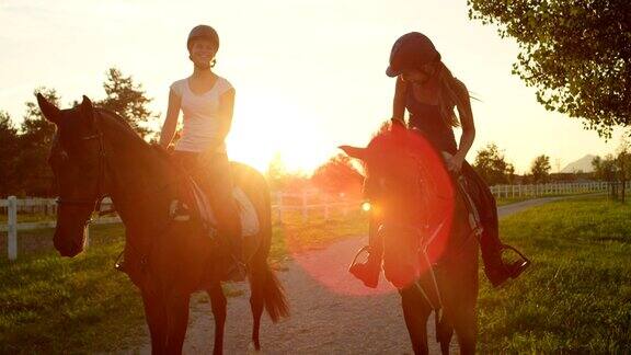 慢镜头:两个笑着的朋友在金色的夕阳下骑着棕色的马