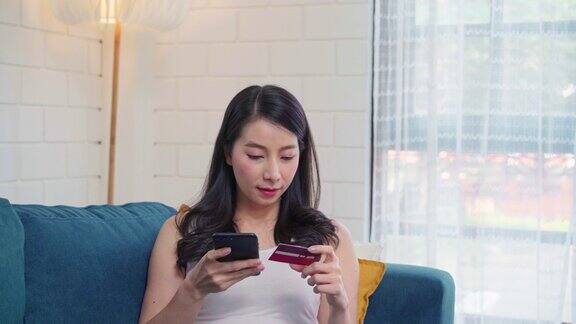 年轻微笑的亚洲女子使用智能手机在网上购物当躺在沙发上在家客厅放松时用信用卡购物居家女性的生活方式概念