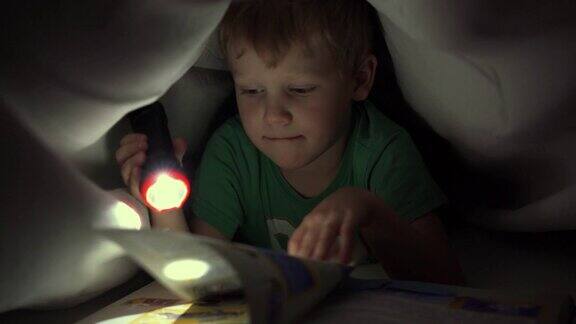 小男孩晚上在毯子下用手电筒看书