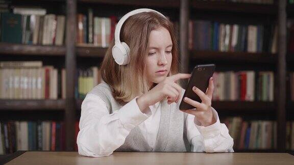 可爱的白人小女孩拿着智能手机玩手机应用坐在图书馆里玩游戏孩子在手机里学习看视频玩手机技术