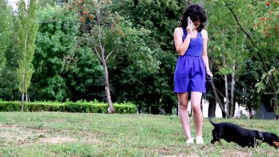一个年轻女子和她的腊肠小狗站在公园里正在讲手机