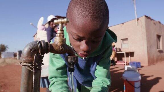 在南非由于严重的干旱年轻的非洲男孩从水龙头喝水而妇女排队收集塑料容器中的水