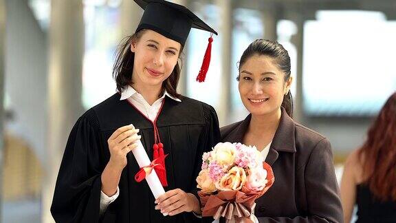 美丽的微笑毕业的学生女孩的肖像年轻的女人和她的母亲在帽子长袍走和拿着文凭