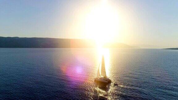 航拍长镜头一艘航行的双体游艇与升起的帆旅行通过公海阳光照耀着海岸边的山丘