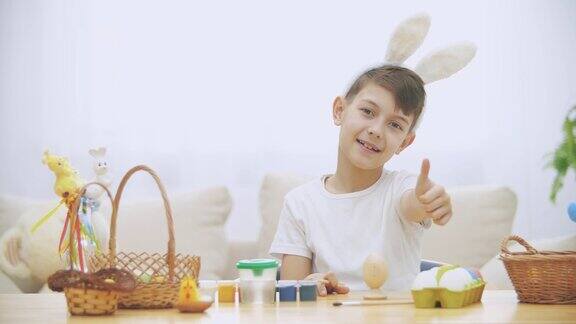 微笑的男孩正在用画笔给复活节彩蛋上色戴着兔耳朵然后竖起左手拇指