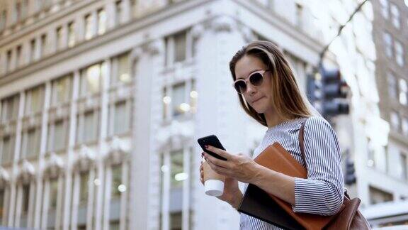 年轻忙碌的女商人戴着太阳镜拿着文件用着智能手机在街上喝着咖啡