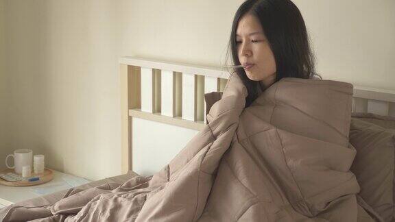 生病的亚洲妇女坐在床上盖着毯子有寒冷颤抖用温度计测量体温看着摄像机