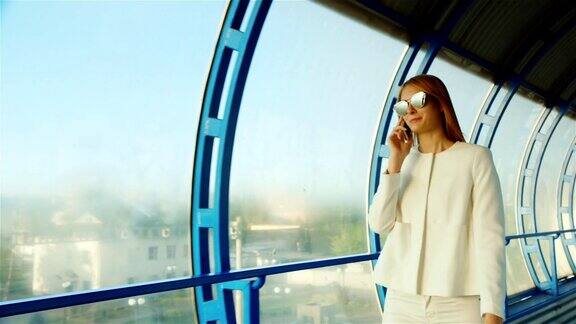 穿着白色西装戴着墨镜的女商人在玻璃窗户的背景下使用手机