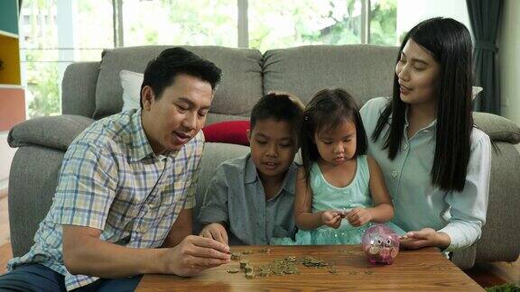 亚洲家庭母亲父亲和两个孩子儿子女儿一起在家里的客厅里用储蓄罐存钱省钱家庭理念