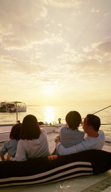 后视图豪华亚洲家庭航行在游艇上坐在蹦床上彼此拥抱享受美丽的海景在日落年轻的父母和年幼的儿子正在欣赏日落时美丽的海景