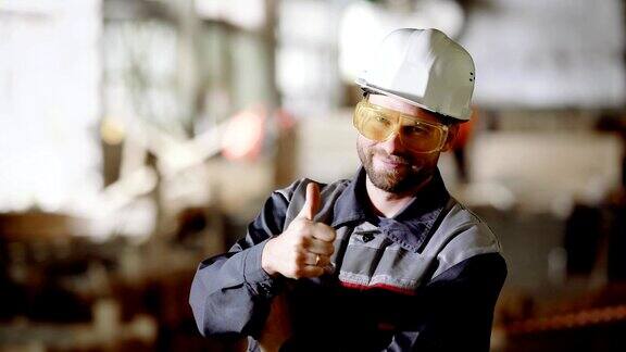近距离的工作人员的制服保护安全帽和眼镜站在施工区域和看镜头一个年轻人在建筑工地工作微笑着竖起大拇指