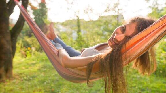 快乐的年轻女子在后院树下的吊床上荡秋千对着镜头微笑