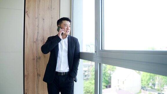 笑容满面的亚洲商人穿着正装在现代办公中心用手机聊天与同伴分享好消息