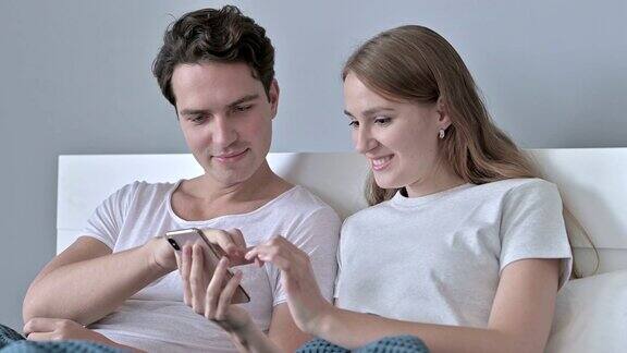 欢快的夫妇坐在床上使用智能手机的肖像