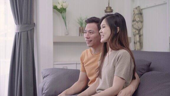亚洲夫妇在家里的客厅看电视喝热咖啡甜蜜的夫妇在家里放松时躺在沙发上享受爱的时刻生活方式夫妇在家里放松