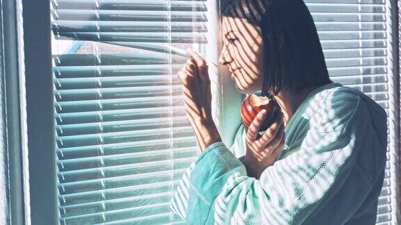 微笑的年轻女子透过百叶窗看着窗外吃着苹果