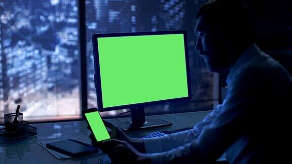 深夜商人在他的私人办公室里用绿色模拟屏幕的个人电脑和智能手机工作可以看到大城市的窗户
