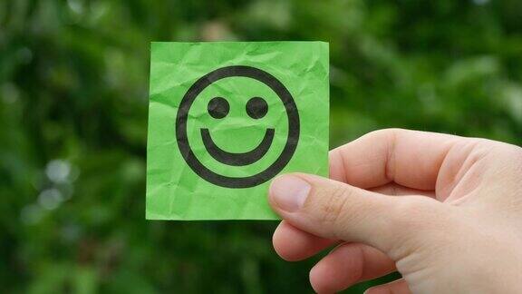 一个女人手里拿着一张绿色的便利贴背景是树叶上面写着一张幸福的脸