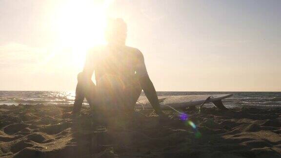 年轻的冲浪者穿着潜水服坐在冲浪板上躺在沙滩上听着音乐跳着舞看着日落时的大海