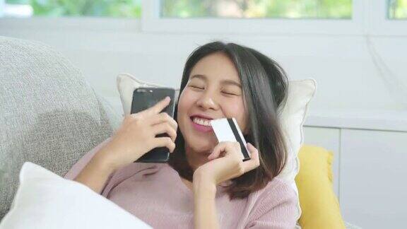 年轻微笑的亚洲女子使用智能手机在网上购物当躺在沙发上在家客厅放松时用信用卡购物生活方式拉丁和西班牙裔妇女在家里