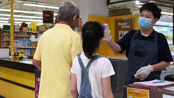 一名亚洲华人中年妇女店主在进入超市前测量老年男子前额测量体温作为新冠肺炎安全预防的标准操作程序