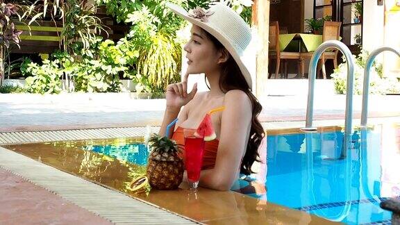 夏天性感的亚洲女人在橙色比基尼与热带饮料在游泳池倾斜了